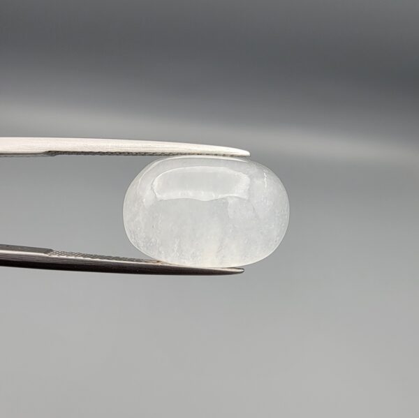 Macro photograph of a 15.34-carat natural Durr-e-Najaf (Clear Rock Crystal Quartz) cabochon held horizontally in stainless steel gem tweezers against a neutral grey background, displaying a smooth oval dome, water-clear transparency, and subtle organic internal veil inclusions.