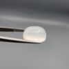 Macro photograph of a 15.34-carat natural Durr-e-Najaf (Clear Rock Crystal Quartz) cabochon held horizontally in stainless steel gem tweezers against a neutral grey background, displaying a smooth oval dome, water-clear transparency, and subtle organic internal veil inclusions.