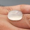 Macro photograph of a 15.34-carat natural Durr-e-Najaf (Clear Rock Crystal Quartz) cabochon held horizontally in stainless steel gem tweezers against a neutral grey background, displaying a smooth oval dome, water-clear transparency, and subtle organic internal veil inclusions.