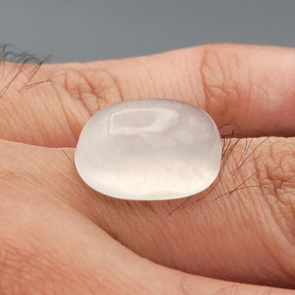Macro photograph of a 15.34-carat natural Durr-e-Najaf (Clear Rock Crystal Quartz) cabochon held horizontally in stainless steel gem tweezers against a neutral grey background, displaying a smooth oval dome, water-clear transparency, and subtle organic internal veil inclusions.