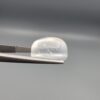 Macro photograph of an 11.36-carat natural Durr-e-Najaf (Clear Rock Crystal Quartz) cabochon held horizontally in stainless steel gem tweezers against a neutral grey background, displaying a smooth oval dome, water-clear transparency, and subtle organic internal veil inclusions.