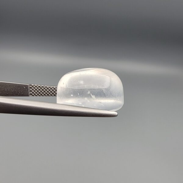 Macro photograph of an 11.36-carat natural Durr-e-Najaf (Clear Rock Crystal Quartz) cabochon held horizontally in stainless steel gem tweezers against a neutral grey background, displaying a smooth oval dome, water-clear transparency, and subtle organic internal veil inclusions.