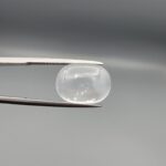Macro photograph of an 11.36-carat natural Durr-e-Najaf (Clear Rock Crystal Quartz) cabochon held horizontally in stainless steel gem tweezers against a neutral grey background, displaying a smooth oval dome, water-clear transparency, and subtle organic internal veil inclusions.