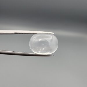 Macro photograph of an 11.36-carat natural Durr-e-Najaf (Clear Rock Crystal Quartz) cabochon held horizontally in stainless steel gem tweezers against a neutral grey background, displaying a smooth oval dome, water-clear transparency, and subtle organic internal veil inclusions.