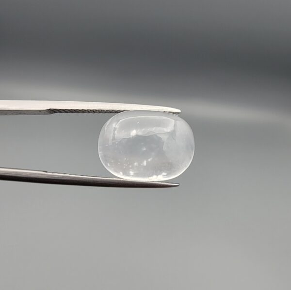 Macro photograph of an 11.36-carat natural Durr-e-Najaf (Clear Rock Crystal Quartz) cabochon held horizontally in stainless steel gem tweezers against a neutral grey background, displaying a smooth oval dome, water-clear transparency, and subtle organic internal veil inclusions.