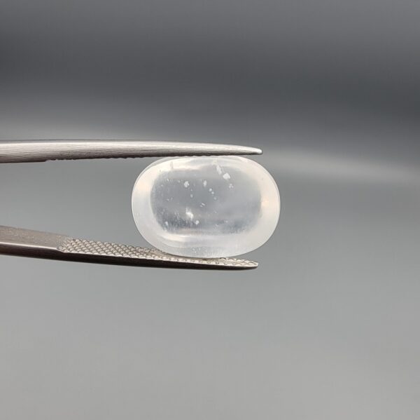 Macro photograph of an 11.36-carat natural Durr-e-Najaf (Clear Rock Crystal Quartz) cabochon held horizontally in stainless steel gem tweezers against a neutral grey background, displaying a smooth oval dome, water-clear transparency, and subtle organic internal veil inclusions.