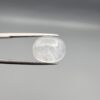 Macro photograph of a 10.46-carat natural Durr-e-Najaf (Clear Rock Crystal Quartz) cabochon held horizontally in stainless steel gem tweezers against a neutral grey background, displaying a smooth oval dome, water-clear transparency, and subtle organic internal veil inclusions.