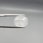 Macro photograph of a 10.46-carat natural Durr-e-Najaf (Clear Rock Crystal Quartz) cabochon held horizontally in stainless steel gem tweezers against a neutral grey background, displaying a smooth oval dome, water-clear transparency, and subtle organic internal veil inclusions.