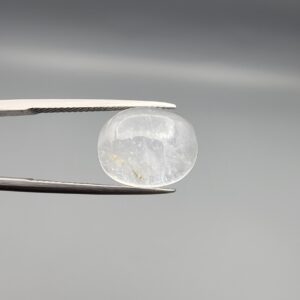 Macro photograph of a 10.46-carat natural Durr-e-Najaf (Clear Rock Crystal Quartz) cabochon held horizontally in stainless steel gem tweezers against a neutral grey background, displaying a smooth oval dome, water-clear transparency, and subtle organic internal veil inclusions.