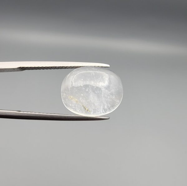 Macro photograph of a 10.46-carat natural Durr-e-Najaf (Clear Rock Crystal Quartz) cabochon held horizontally in stainless steel gem tweezers against a neutral grey background, displaying a smooth oval dome, water-clear transparency, and subtle organic internal veil inclusions.