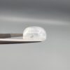 Macro photograph of a 10.46-carat natural Durr-e-Najaf (Clear Rock Crystal Quartz) cabochon held horizontally in stainless steel gem tweezers against a neutral grey background, displaying a smooth oval dome, water-clear transparency, and subtle organic internal veil inclusions.