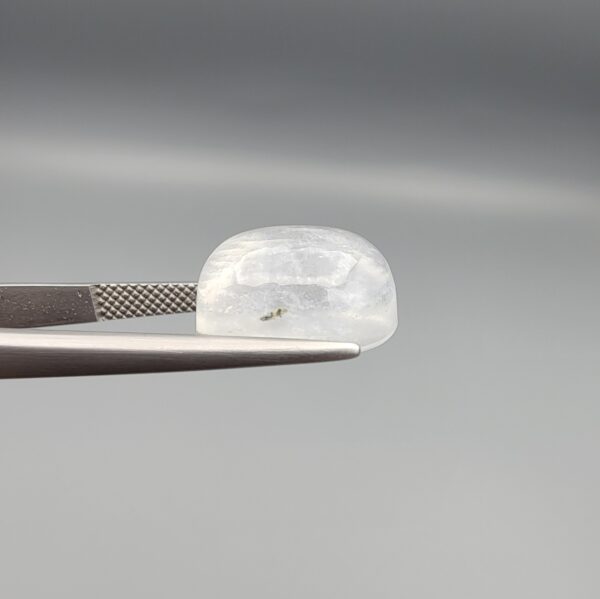Macro photograph of a 10.46-carat natural Durr-e-Najaf (Clear Rock Crystal Quartz) cabochon held horizontally in stainless steel gem tweezers against a neutral grey background, displaying a smooth oval dome, water-clear transparency, and subtle organic internal veil inclusions.