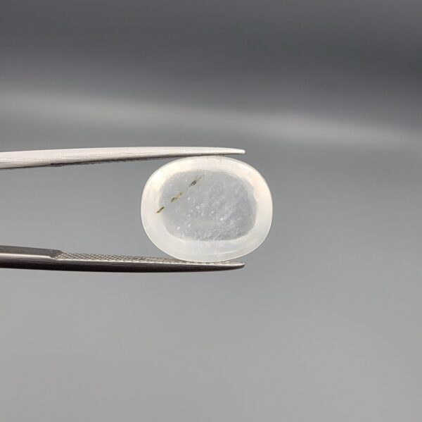Macro photograph of a 10.46-carat natural Durr-e-Najaf (Clear Rock Crystal Quartz) cabochon held horizontally in stainless steel gem tweezers against a neutral grey background, displaying a smooth oval dome, water-clear transparency, and subtle organic internal veil inclusions.