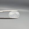 Macro photograph of a 9.24-carat natural Durr-e-Najaf (Clear Rock Crystal Quartz) cabochon held horizontally in stainless steel gem tweezers against a neutral grey background, displaying a smooth oval dome, water-clear transparency, and subtle organic internal veil inclusions.