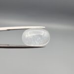Macro photograph of a 9.24-carat natural Durr-e-Najaf (Clear Rock Crystal Quartz) cabochon held horizontally in stainless steel gem tweezers against a neutral grey background, displaying a smooth oval dome, water-clear transparency, and subtle organic internal veil inclusions.