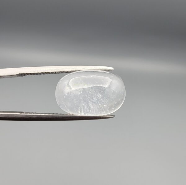 Macro photograph of a 9.24-carat natural Durr-e-Najaf (Clear Rock Crystal Quartz) cabochon held horizontally in stainless steel gem tweezers against a neutral grey background, displaying a smooth oval dome, water-clear transparency, and subtle organic internal veil inclusions.
