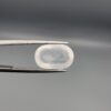 Macro photograph of a 9.24-carat natural Durr-e-Najaf (Clear Rock Crystal Quartz) cabochon held horizontally in stainless steel gem tweezers against a neutral grey background, displaying a smooth oval dome, water-clear transparency, and subtle organic internal veil inclusions.