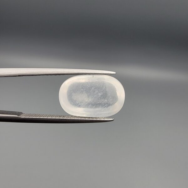 Macro photograph of a 9.24-carat natural Durr-e-Najaf (Clear Rock Crystal Quartz) cabochon held horizontally in stainless steel gem tweezers against a neutral grey background, displaying a smooth oval dome, water-clear transparency, and subtle organic internal veil inclusions.