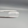 Macro photograph of an 11.47-carat natural Durr-e-Najaf (Clear Rock Crystal Quartz) cabochon held horizontally in stainless steel gem tweezers against a neutral grey background, displaying a smooth oval dome, water-clear transparency, and subtle organic internal veil inclusions.