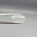 Macro photograph of an 11.47-carat natural Durr-e-Najaf (Clear Rock Crystal Quartz) cabochon held horizontally in stainless steel gem tweezers against a neutral grey background, displaying a smooth oval dome, water-clear transparency, and subtle organic internal veil inclusions.