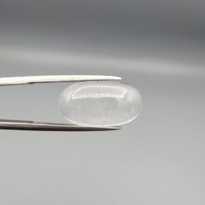 Macro photograph of an 11.47-carat natural Durr-e-Najaf (Clear Rock Crystal Quartz) cabochon held horizontally in stainless steel gem tweezers against a neutral grey background, displaying a smooth oval dome, water-clear transparency, and subtle organic internal veil inclusions.