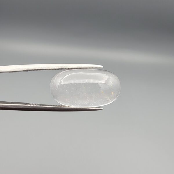 Macro photograph of an 11.47-carat natural Durr-e-Najaf (Clear Rock Crystal Quartz) cabochon held horizontally in stainless steel gem tweezers against a neutral grey background, displaying a smooth oval dome, water-clear transparency, and subtle organic internal veil inclusions.