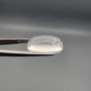 Macro photograph of an 11.47-carat natural Durr-e-Najaf (Clear Rock Crystal Quartz) cabochon held horizontally in stainless steel gem tweezers against a neutral grey background, displaying a smooth oval dome, water-clear transparency, and subtle organic internal veil inclusions.