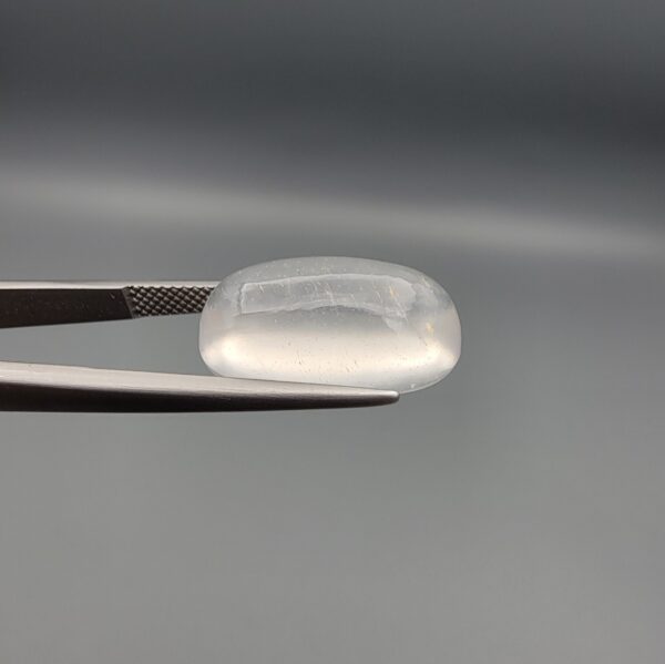 Macro photograph of an 11.47-carat natural Durr-e-Najaf (Clear Rock Crystal Quartz) cabochon held horizontally in stainless steel gem tweezers against a neutral grey background, displaying a smooth oval dome, water-clear transparency, and subtle organic internal veil inclusions.