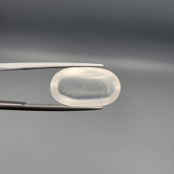 Macro photograph of an 11.47-carat natural Durr-e-Najaf (Clear Rock Crystal Quartz) cabochon held horizontally in stainless steel gem tweezers against a neutral grey background, displaying a smooth oval dome, water-clear transparency, and subtle organic internal veil inclusions.