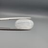 Macro photograph of a 7.95-carat natural Durr-e-Najaf (Clear Rock Crystal Quartz) cabochon held horizontally in stainless steel gem tweezers against a neutral grey background, displaying a smooth oval dome, water-clear transparency, and subtle organic internal veil inclusions.