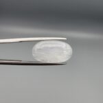 Macro photograph of a 7.95-carat natural Durr-e-Najaf (Clear Rock Crystal Quartz) cabochon held horizontally in stainless steel gem tweezers against a neutral grey background, displaying a smooth oval dome, water-clear transparency, and subtle organic internal veil inclusions.