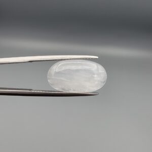 Macro photograph of a 7.95-carat natural Durr-e-Najaf (Clear Rock Crystal Quartz) cabochon held horizontally in stainless steel gem tweezers against a neutral grey background, displaying a smooth oval dome, water-clear transparency, and subtle organic internal veil inclusions.