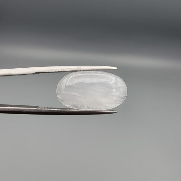 Macro photograph of a 7.95-carat natural Durr-e-Najaf (Clear Rock Crystal Quartz) cabochon held horizontally in stainless steel gem tweezers against a neutral grey background, displaying a smooth oval dome, water-clear transparency, and subtle organic internal veil inclusions.