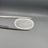 Macro photograph of a 7.95-carat natural Durr-e-Najaf (Clear Rock Crystal Quartz) cabochon held horizontally in stainless steel gem tweezers against a neutral grey background, displaying a smooth oval dome, water-clear transparency, and subtle organic internal veil inclusions.