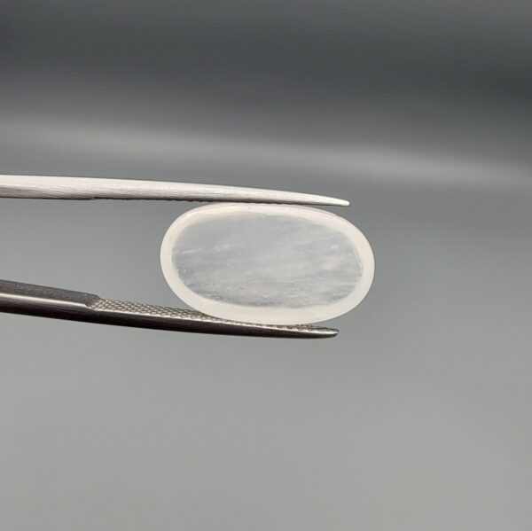 Macro photograph of a 7.95-carat natural Durr-e-Najaf (Clear Rock Crystal Quartz) cabochon held horizontally in stainless steel gem tweezers against a neutral grey background, displaying a smooth oval dome, water-clear transparency, and subtle organic internal veil inclusions.
