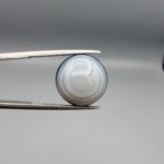 Multi-angle macro photographs of a 12.06-carat natural African Eye Agate (Sulemani Aqeeq) held in gem tweezers, showing a high round cabochon dome with translucent white and grey halo banding, a luminous bright focal center, and a base with natural black mineral plume inclusions.