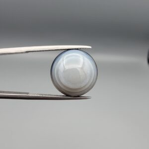 Multi-angle macro photographs of a 12.06-carat natural African Eye Agate (Sulemani Aqeeq) held in gem tweezers, showing a high round cabochon dome with translucent white and grey halo banding, a luminous bright focal center, and a base with natural black mineral plume inclusions.