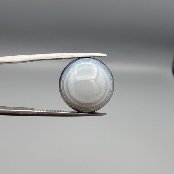 Multi-angle macro photographs of a 12.06-carat natural African Eye Agate (Sulemani Aqeeq) held in gem tweezers, showing a high round cabochon dome with translucent white and grey halo banding, a luminous bright focal center, and a base with natural black mineral plume inclusions.