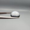 Multi-angle macro photographs of a 12.06-carat natural African Eye Agate (Sulemani Aqeeq) held in gem tweezers, showing a high round cabochon dome with translucent white and grey halo banding, a luminous bright focal center, and a base with natural black mineral plume inclusions.