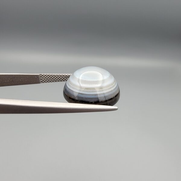 Multi-angle macro photographs of a 12.06-carat natural African Eye Agate (Sulemani Aqeeq) held in gem tweezers, showing a high round cabochon dome with translucent white and grey halo banding, a luminous bright focal center, and a base with natural black mineral plume inclusions.