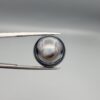Multi-angle macro photographs of a 12.60-carat natural African Eye Agate (Sulemani Aqeeq) held in gem tweezers, showcasing a high round cabochon dome with concentric white, grey, and jet-black banding, a brownish-grey core pupil, and a polished black base with a subtle light halo center.