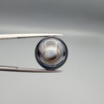Multi-angle macro photographs of a 12.60-carat natural African Eye Agate (Sulemani Aqeeq) held in gem tweezers, showcasing a high round cabochon dome with concentric white, grey, and jet-black banding, a brownish-grey core pupil, and a polished black base with a subtle light halo center.
