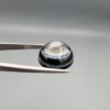 Multi-angle macro photographs of a 12.60-carat natural African Eye Agate (Sulemani Aqeeq) held in gem tweezers, showcasing a high round cabochon dome with concentric white, grey, and jet-black banding, a brownish-grey core pupil, and a polished black base with a subtle light halo center.
