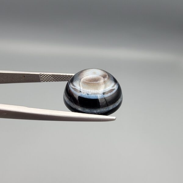Multi-angle macro photographs of a 12.60-carat natural African Eye Agate (Sulemani Aqeeq) held in gem tweezers, showcasing a high round cabochon dome with concentric white, grey, and jet-black banding, a brownish-grey core pupil, and a polished black base with a subtle light halo center.