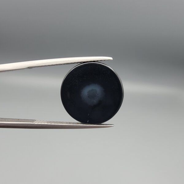 Multi-angle macro photographs of a 12.60-carat natural African Eye Agate (Sulemani Aqeeq) held in gem tweezers, showcasing a high round cabochon dome with concentric white, grey, and jet-black banding, a brownish-grey core pupil, and a polished black base with a subtle light halo center.