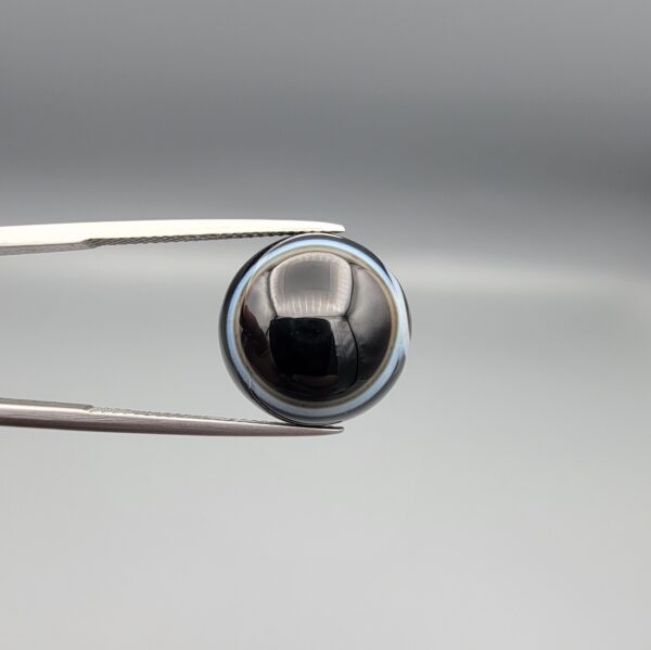 Multi-angle macro photographs of a 14.77-carat natural African Eye Agate (Sulemani Aqeeq) held in gem tweezers, showing a high round cabochon dome with a deep onyx-black core, sharp white outer ringing, tiered side profile bands, and a flat jet-black base with fine white agate pinstripes.
