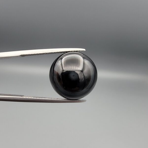 Multi-angle macro photographs of a 12.99-carat natural African Eye Agate (Sulemani Aqeeq) held in gem tweezers, showcasing a high round cabochon dome with deep charcoal-black body color, subtle flank fortification banding, and a base filled with intricate dendritic mineral inclusions.