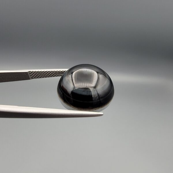 Multi-angle macro photographs of a 12.99-carat natural African Eye Agate (Sulemani Aqeeq) held in gem tweezers, showcasing a high round cabochon dome with deep charcoal-black body color, subtle flank fortification banding, and a base filled with intricate dendritic mineral inclusions.