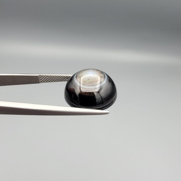 Multi-angle macro photographs of a 13.29-carat natural African Banded Eye Agate (Sulemani Aqeeq) held in gem tweezers, displaying a high round cabochon dome with concentric white and grey ringing around a translucent light core, a black border, and a reverse side showing undulating blue-grey wave banding.