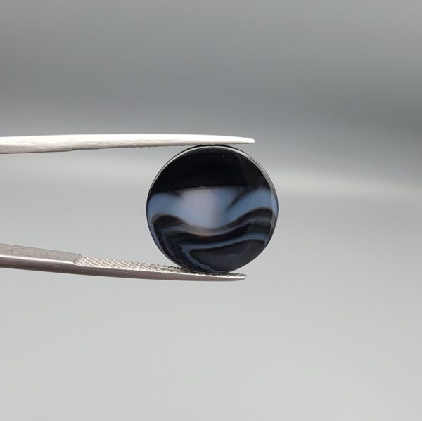 Multi-angle macro photographs of a 13.29-carat natural African Banded Eye Agate (Sulemani Aqeeq) held in gem tweezers, displaying a high round cabochon dome with concentric white and grey ringing around a translucent light core, a black border, and a reverse side showing undulating blue-grey wave banding.